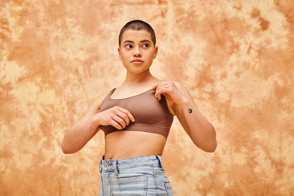 body positivity movement, curvy young woman with tattoos posing in jeans and crop top on mottled beige background, representation of body, confidence, casual attire, generation z, looking away