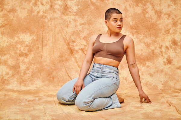 body positivity, denim fashion, curvy and tattooed woman in jeans and crop top sitting on mottled beige background, casual attire, looking away, self-acceptance, generation z, body diversity 