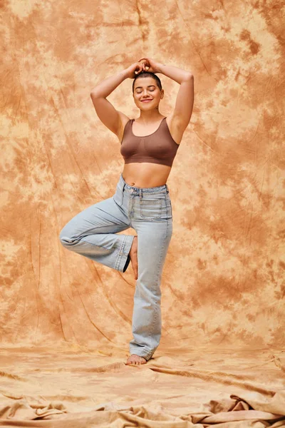 积极的体态 自信的 卷曲的 快乐的女人在庄稼顶上 牛仔裤在斑驳的米色背景上 瑜伽姿势 自我接纳 Z世代 牛仔时尚 — 图库照片