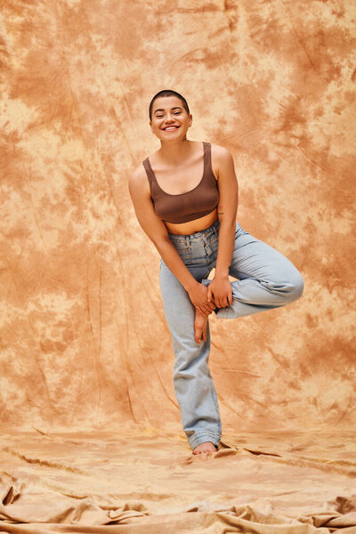 body positivity and confidence, happy young woman in crop top and jeans posing on mottled beige background, casual attire, self-acceptance, generation z, tattooed, smile, full length, denim fashion 