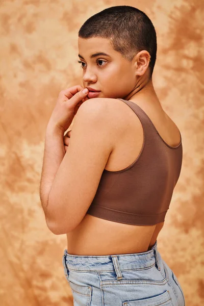 stock image representation of body, curvy and young woman in crop top and jeans posing with hand near lips on mottled beige background, short haired, self-acceptance, generation z, tattooed, different shapes 