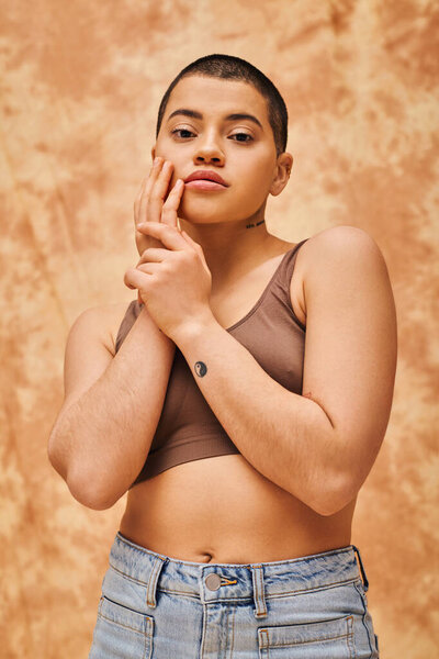 body positivity and confidence, curvy and young woman in crop top and jeans posing on mottled beige background, short haired, self-acceptance, generation z, tattooed, different shapes 