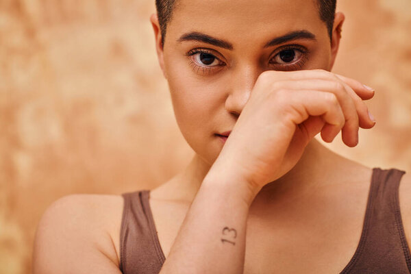 natural look, self-acceptance, tattooed young woman with short hair posing on mottled beige background, hand near face, individuality, modern generation z, beauty, looking at camera 