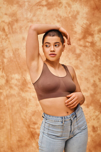body love, curvy young woman in crop top posing with raised hand on mottled beige background, looking away, representation of body, different shapes, generation z, youth, tattooed, relaxed pose 