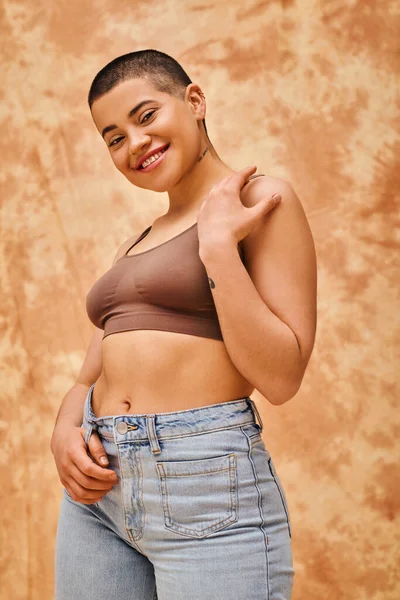 body love, jeans look, curvy and tattooed woman in casual attire standing on mottled beige background, confidence, self-acceptance, generation z, body diversity, pretty and positive