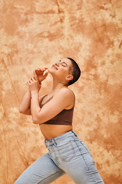 denim fashion, gen z, happy curvy woman with tattoo posing on med lead beige fone, body positivity movement, self-esteem, confidence, short haired model, youth culture 