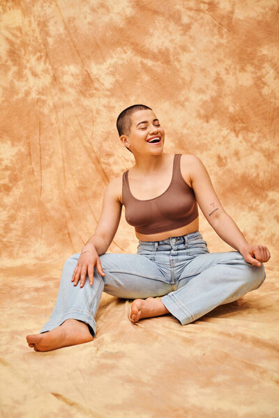 self acceptance, amazed and tattooed woman in jeans and crop top sitting on mottled beige background, casual attire, looking away, self-acceptance, generation z, body diversity, laughter 