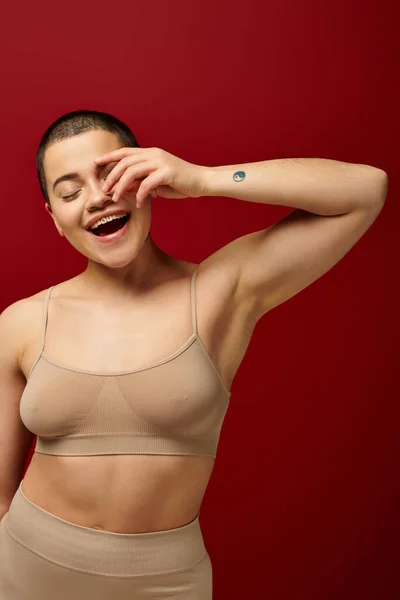 self-esteem, excited and tattooed woman in beige underwear posing on red background, body positivity, curvy fashion, comfortable in skin, self-acceptance, gen z, body diversity, hand near face