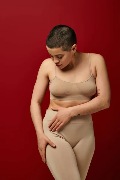 body positive, self-esteem, young woman with short hair and tattoo posing on burgundy background, dark red, curvy fashion, comfortable in skin, female underwear, fashion model, body type