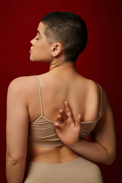 body positive, self-esteem, tattooed young woman with short hair and tattoo posing with hand behind back on burgundy background, dark red, curvy fashion, comfortable in skin, female underwear