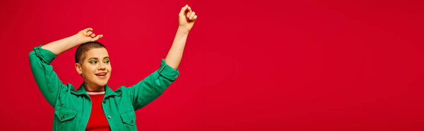 мода и стиль, возбужденная и коротковолосая женщина в зеленом наряде, позирующая с поднятыми руками на красном фоне, поколение Z, молодежная культура, современный бэккап, индивидуальность, личный стиль, баннер 