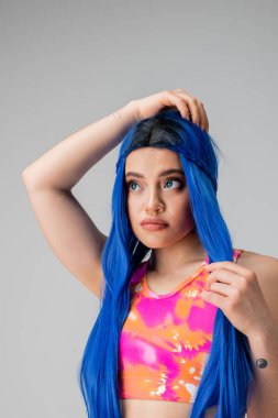 Kendini ifade etme, mavi saçı ayarlayan ve gri arka plana bakan genç bir kadın, izole edilmiş, moda tercihleri, şık görünüş, renkli kıyafetler, günlük kıyafetler, Z kuşağı modası, uzun saç.