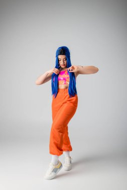 Moda trendleri, renkli mavi saçlı, gri arka planda renkli pozlar veren dövmeli genç kadın, tam boy, funky görünüm, bireysellik, modern tarz, kentsel moda, canlı renk, model. 