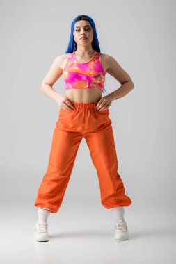 Moda, dövmeli, mavi saçlı, elleri kalçasında poz veren, gri arka planda renkli kıyafetler giyen, tam boy, bireysellik, modern tarz, şehir modası, canlı renk.