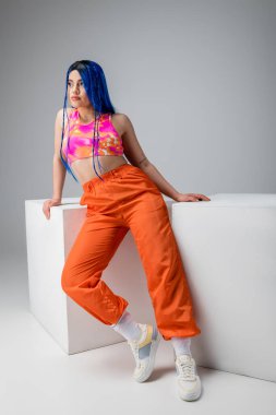 Moda trendleri, mavi saçlı, gri arka planda beyaz küplerin yanında renkli pozlar veren dövmeli genç kadın, tam boy, bireysellik, modern tarz, şehir tarzı, asi tarzı.