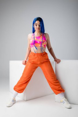 Moda ileri, mavi saçlı, gri arka planda beyaz küplerin yanında duran renkli elbiseli genç kadın, tam boy, bireysellik, modern tarz, kentsel moda, canlı renk, model 