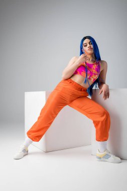 Modern alt kültür, mavi saçlı, gri arka planda beyaz küplerin yanında renkli pozlar veren dövmeli kadın, tam boy, bireysellik, modern tarz, kentsel moda, canlı renk, genç model. 