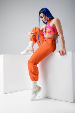 Moda trendleri, mavi saçlı dövmeli genç kadın gri arka planda beyaz küplerle otururken, tam boy, bireysellik, modern tarz, kentsel moda, canlı renk 