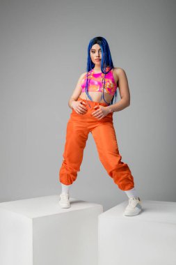 Moda trendi, mavi saçlı genç bir kadın gri arka planda renkli pozlar veriyor, küplerin üzerinde duruyor, tam boy, bireysellik, modern tarz, şehir modası, canlı renk 
