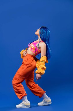 full length, woman with dyed hair, fashion forward, female model with blue hair posing in puffer jacket and orange pants on blue background, vibrant color, urban fashion, individualism  clipart