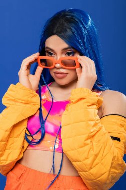 Moda ve tarz, mavi saçlı, turuncu güneş gözlüğü takan genç bir kadın model mavi arka planda izole edilmiş, Z nesli, asi tarzı, renkli kıyafetler, bireysellik, modern kadın. 