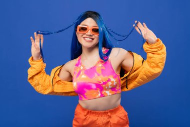 Moda anlayışı, mavi saçlı ve modaya uygun güneş gözlüklü neşeli kadın modeli mavi arka planda izole edilmiş Z jenerasyonu, asi tarzı, renkli kıyafetler, bireysellik, modern pozitif kadın. 