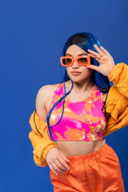 Moda anlayışı, mavi saçlı, modaya uygun güneş gözlüklü genç bir kadın model mavi arka planda izole edilmiş, Z nesli, asi tarzı, renkli kıyafetler, bireysellik, modern kadın. 
