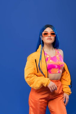 Gen z, moda anlayışı, mavi saçlı genç bayan model, modaya uygun güneş gözlüğü, mavi arka planda izole edilmiş, asi tarzı, renkli kıyafetler, bireysellik, modern kadın. 