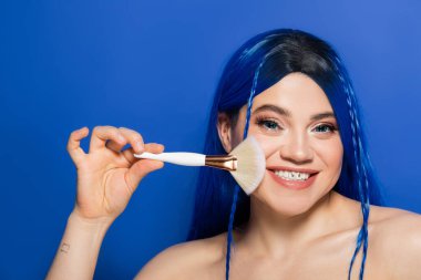 Güzellik trendleri, bireysellik, canlı gözleri ve saçlarıyla kameraya bakan neşeli genç kadın mavi arka planda makyaj fırçası tutarken, kozmetik, kendini ifade etme, yüz ifadesi, gençlik