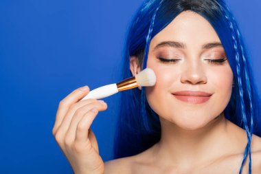 Bireycilik, canlı gözleri ve saçları olan genç bir kadının mavi arka planda kozmetik fırçayı tutarken kameraya bakması, makyaj, kendini ifade etme, yüz ifadesi, gençlik, güzellik eğilimleri 