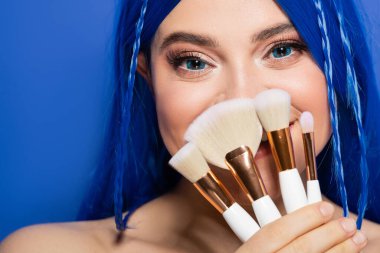 Makyaj aletleri, parlayan cilt, parlak saçlı ve mavi arka planda kozmetik fırça tutan kadın model, makyaj, güzellik trendleri, yüz ifadesi, güzellik endüstrisi, yüz kaplaması 