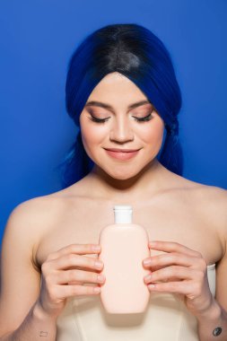 Saç bakımı konsepti, canlı saç renginde portresi mavi arka planda çıplak omuz pozu, şampuanlı kozmetik şişe, güzellik trendleri. 