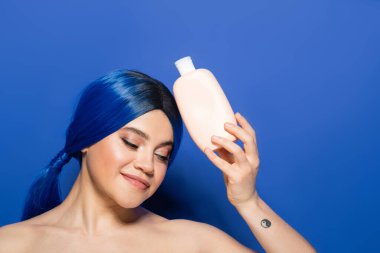 Güzellik trendleri, vücut ve saç bakımı konsepti, dövmeli genç kadın portresi parlak saç renginde mavi arka planda çıplak omuzlu poz, şampuanlı kozmetik şişe, reklam 
