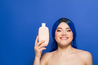 Güzellik trendleri, saç bakımı konsepti, parlak saçlı mutlu bir kadının portresi mavi arka planda çıplak omuzlarıyla poz vermesi, şampuanlı kozmetik bir şişe tutması, reklam. 