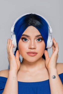 Müzik aşığı, mavi saçlı genç bir kadın gri arka planda kablosuz kulaklıkla müzik dinliyor, enerjik gençlik, bireysellik, modern altkültür, kendini ifade etme, dövme, ses 
