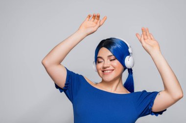 Kendini ifade eden, mavi saçlı neşeli genç bir kadın gri arka planda kablosuz kulaklıkla müzik dinliyor, dans ediyor, hayat dolu gençlik, bireysellik, modern altkültür, dövme, ses 