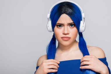 Kendini ifade etme, mavi saçlı hoşnutsuz genç kadın gri arka planda kablosuz kulaklıkla müzik dinliyor, enerjik gençlik, bireysellik, modern altkültür, ses, kameraya bakıyor.  