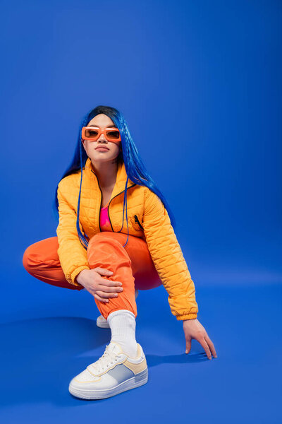 individualism, full length of pretty female model with blue hair and trendy sunglasses sitting on haunches on blue background, rebel style, modern fashion, trendy accessory, generation z, 