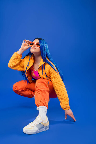 generation z, full length of pretty female model with blue hair and trendy sunglasses sitting on haunches on blue background, rebel style, modern fashion, trendy accessory, gen z