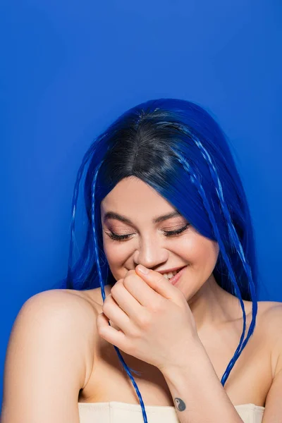 vibrant youth, self expression, portrait of shy young woman with dyed hair smiling and covering mouth on blue background, hair color, individualism, female model with makeup and trendy hairstyle