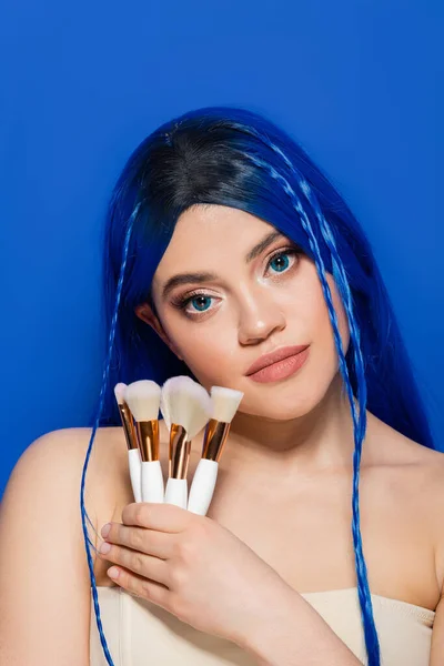 beauty industry, individualism, young woman with vibrant hair and eyes looking at camera while holding cosmetic brushes on blue background, makeup, beauty trends, visage, youth, self expression