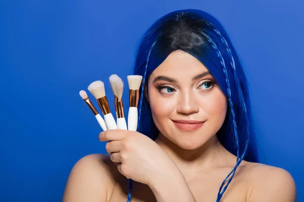 beauty industry, individualism, cheerful young woman with vibrant hair and eyes looking away while holding cosmetic brushes on blue background, makeup, beauty trends, visage, self expression