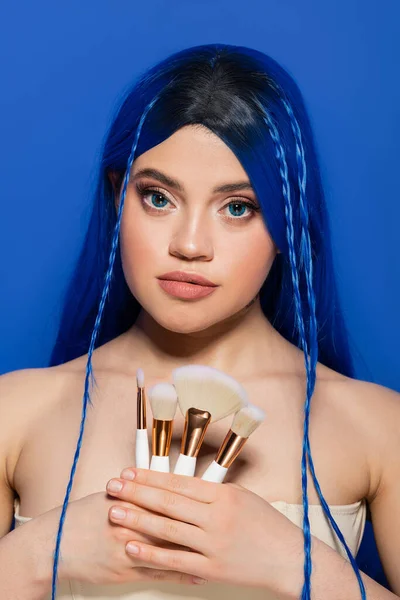 beauty industry, individualism, young woman with vibrant hair and eyes looking at camera while holding makeup brushes on blue background, cosmetic, beauty trends, visage, youth, self expression