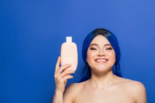 beauty trends, hair care concept, portrait of happy young woman with vibrant hair color posing with bare shoulders on blue background, holding cosmetic bottle with shampoo, advertisement