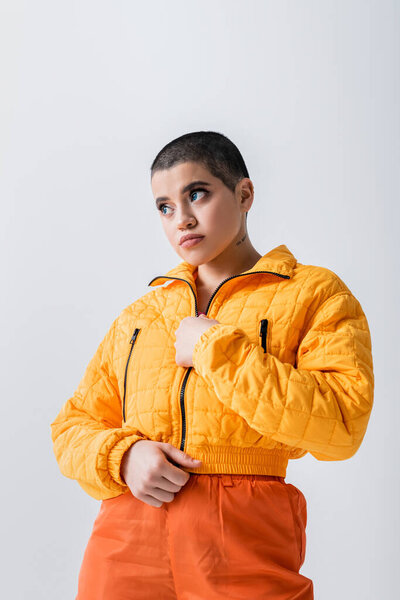 outerwear, tattooed and young woman with short hair zipping yellow puffer jacket on grey background, urban fashion, vibrant youth, trendy outfit, stylish look, studio photography, modern attire 