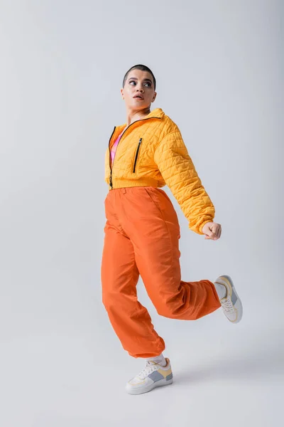 stylish look, outerwear, casual attire, fashion model posing in yellow puffer jacket and orange pants on grey background, woman with short hair running and looking away, modern subculture