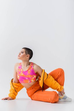 enerjik gençlik, dövmeli kısa saçlı genç kadın gri arka planda oturuyor, z nesli, moda ileri, renkli kıyafetler, kadın modeli kişisel tarzı, kendini ifade etme, moda kıyafeti 