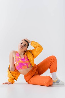 canlı renkler, kısa saçlı dövmeli genç bir kadın gri arka planda oturuyor, z nesli, moda ileri, renkli kıyafetler, kadın modeli, kişisel tarzı, kendini ifade etme, moda kıyafeti 