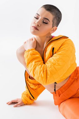 Canlı renkler, kısa saçlı, gri arka planda oturan dövmeli genç bir kadın, z jenerasyonu, ileri moda, renkli kıyafetler, kapalı gözlü kadın modeli, kendini ifade etme, moda kıyafeti. 