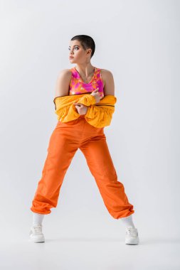 personal style, warm clothing, fashionable young woman with short hair posing in puffer jacket and vibrant pants on grey background, outerwear, modern subculture, youthful energy, full length 
